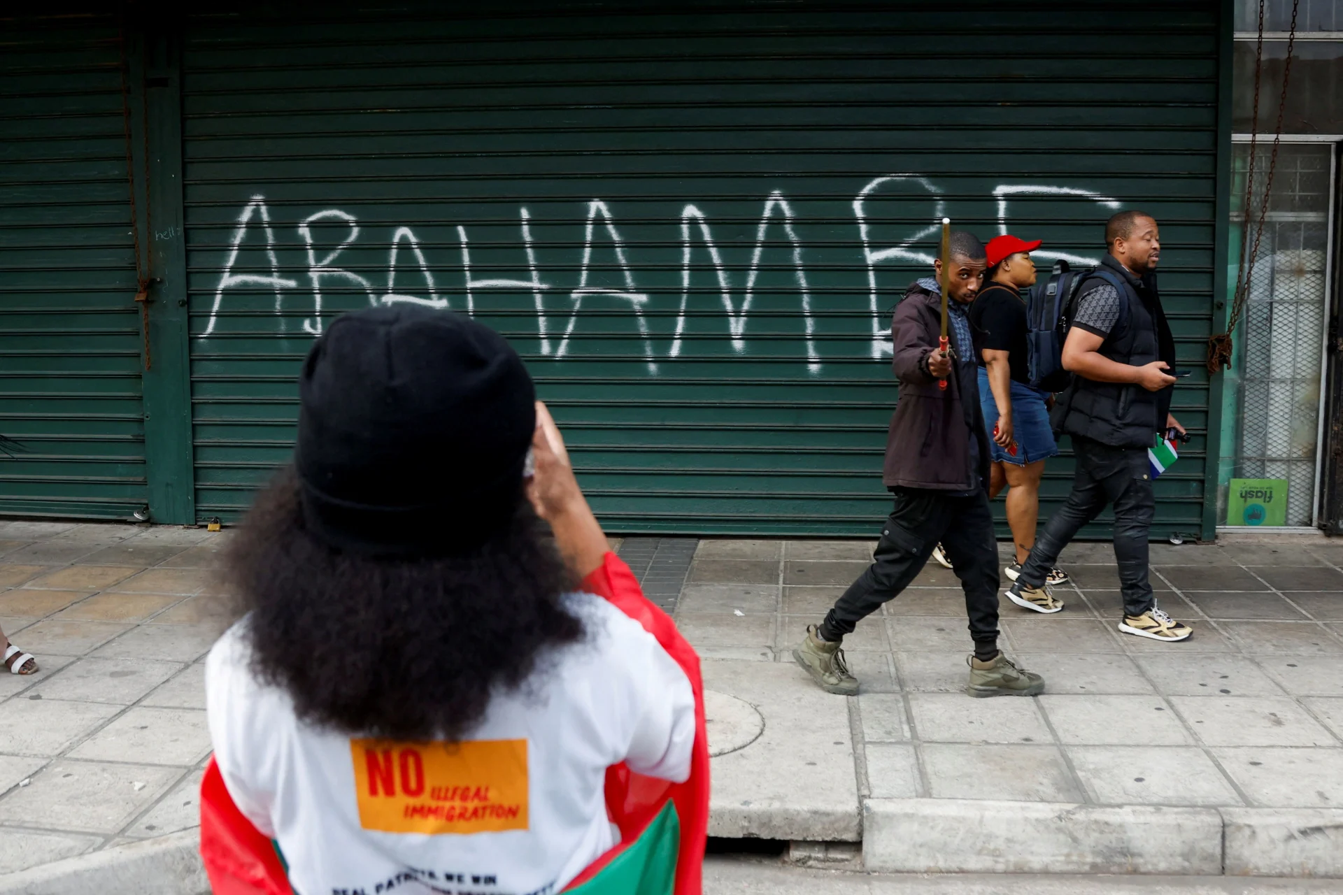People walk past a slogan reading, “They must go,” written on the shutter of a shop as anti-immigrant protesters march on the day of an unofficial deadline set by anti-immigrant groups for all undocumented migrants to leave, in Durban, South Africa, on June 30, 2026. Reuters/Rogan Ward